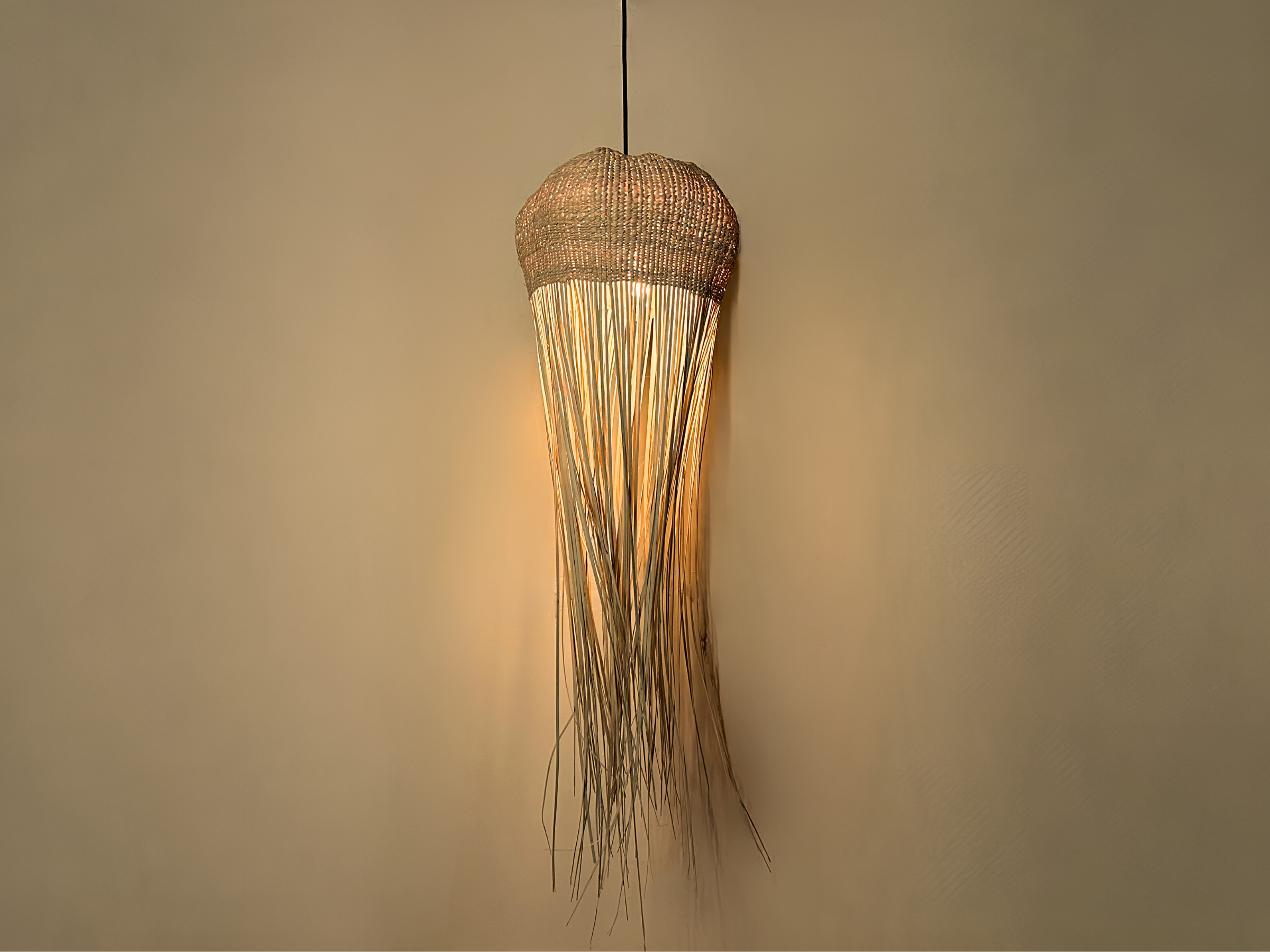 Tall jellyfish-inspired pendant light with woven top and flowing raffia strands, softly glowing against a neutral wall.