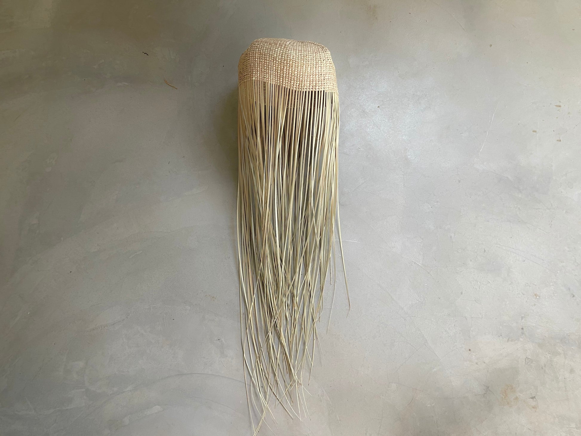 Tall jellyfish-inspired pendant light with woven top and flowing raffia strands, lying on a concrete floor
