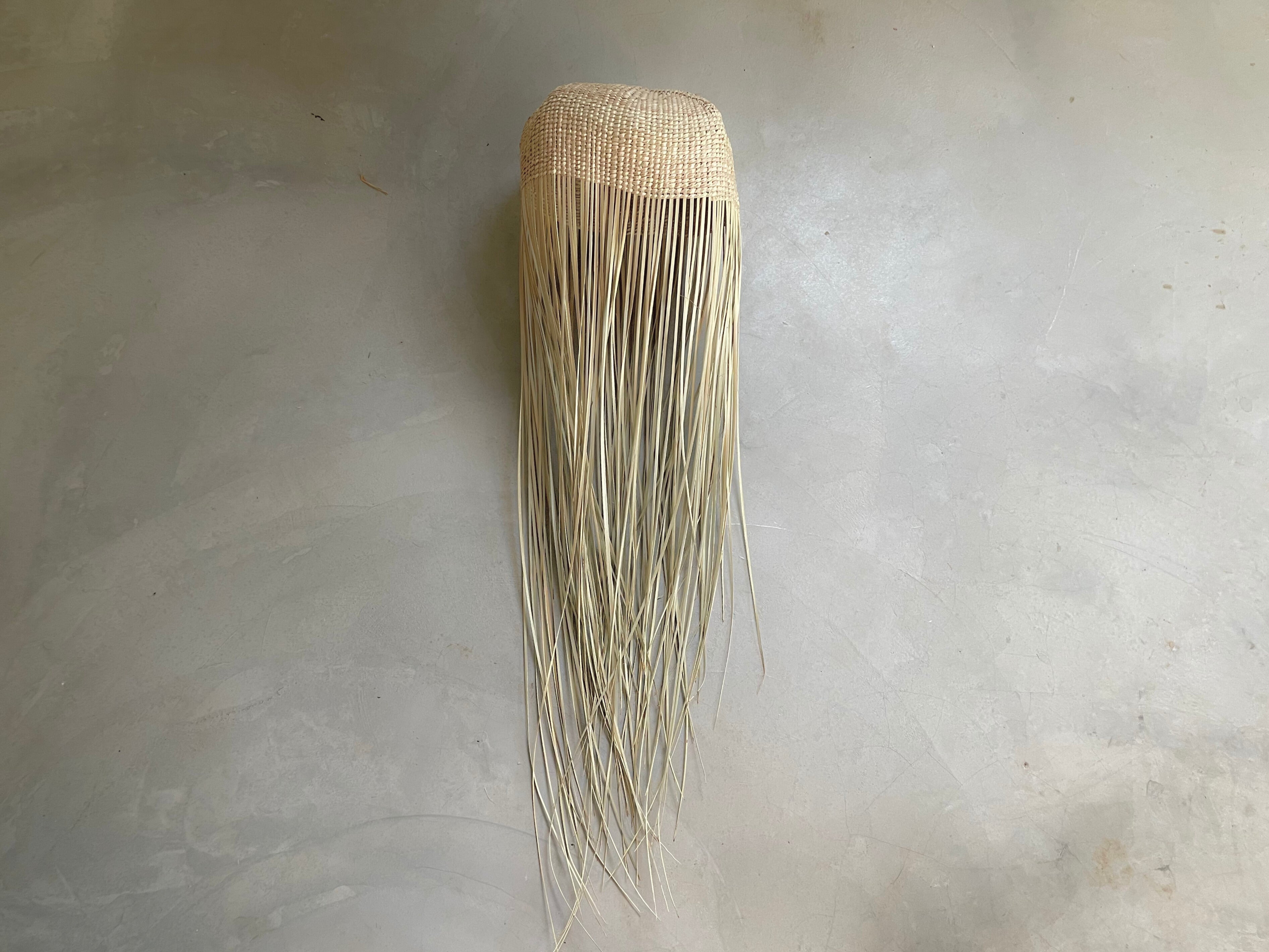 Tall jellyfish-inspired pendant light with woven top and flowing raffia strands, lying on a concrete floor